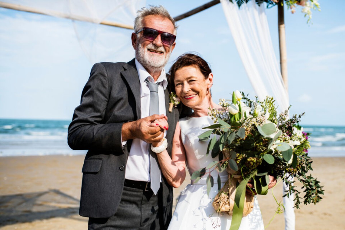 Un couple marié mature se tenant la main sur une plage, entouré d’une arche florale, souriant avec complicité lors de la célébration de leurs 39 ans de mariage – noces de crêpes